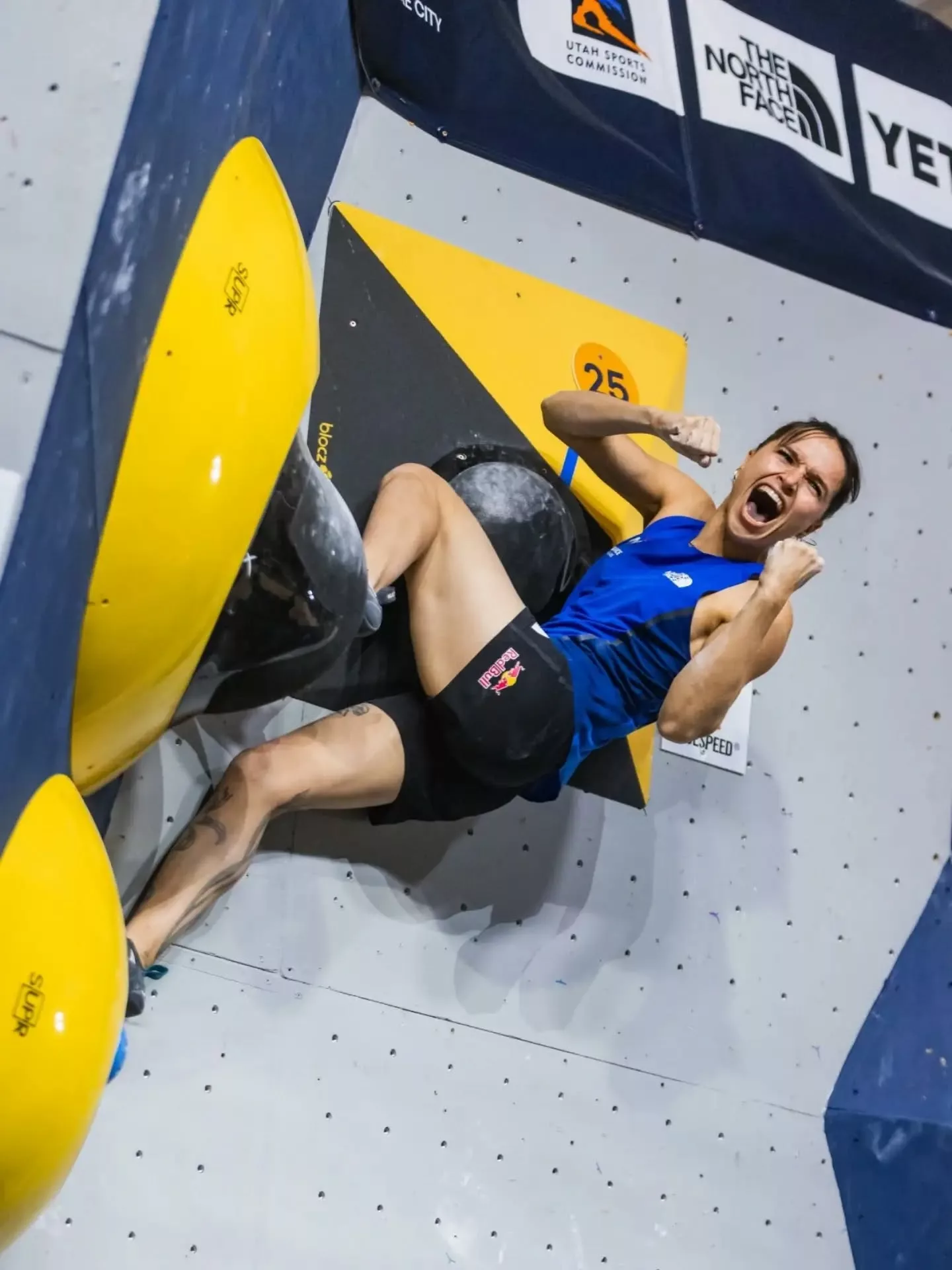 Oriane Bertone sur un bloc d’escalade à Salt Lake City, en maillot bleu, le poing levé près des prises jaunes.