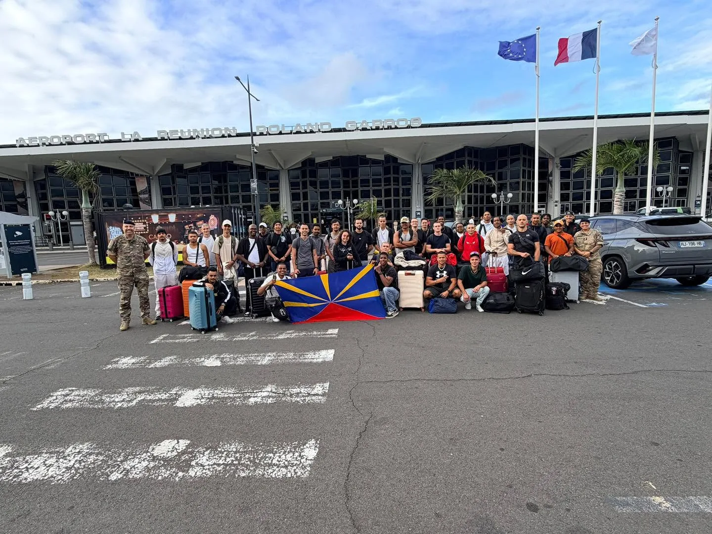 Un groupe de candidats et d’accompagnateurs avec des valises et un drapeau réunionnais devant l’aéroport Roland-Garros.
