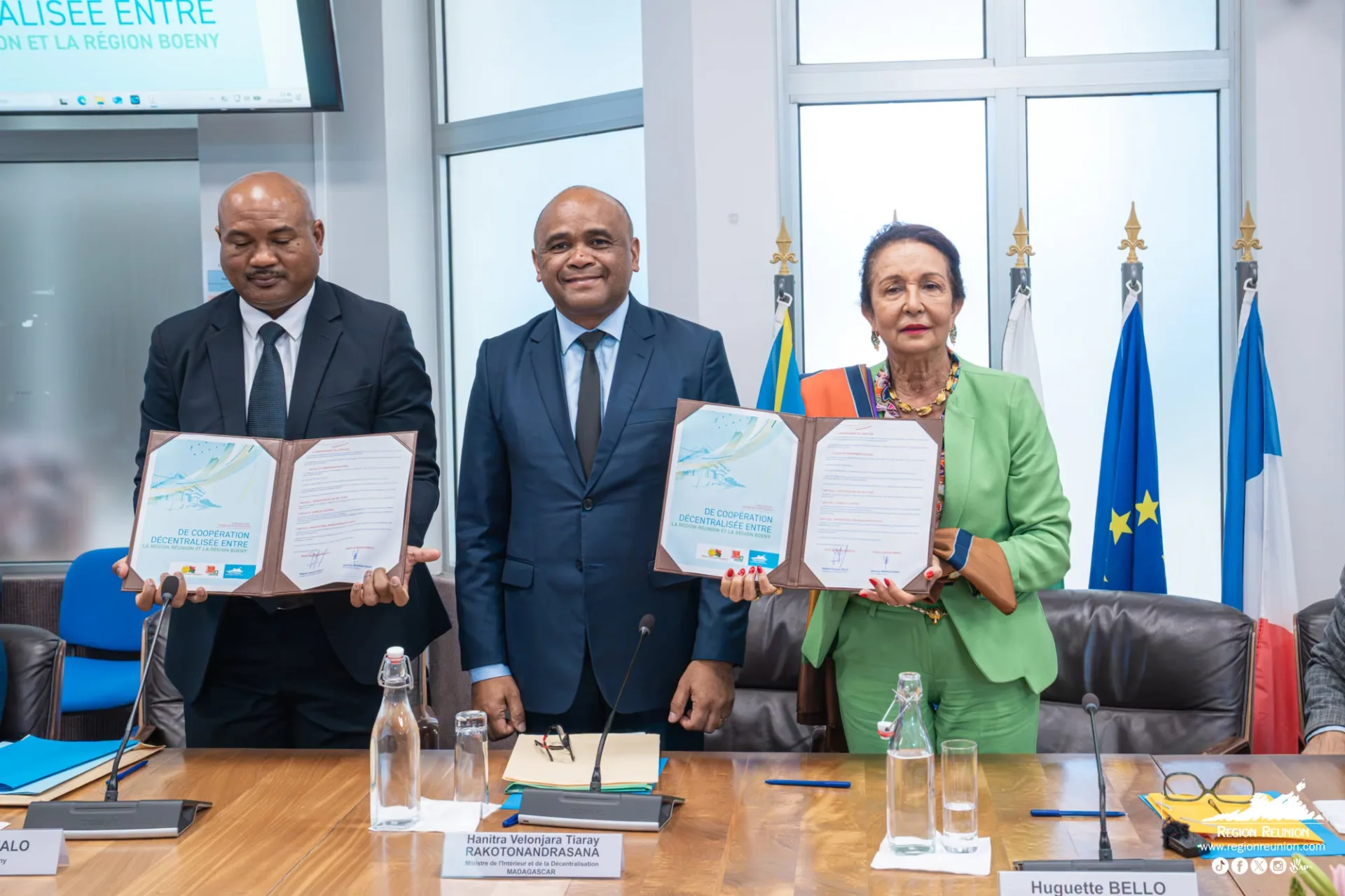 Trois représentants présentent les documents de l’accord de coopération Réunion–Boeny, devant les drapeaux à l’Hôtel de Région.